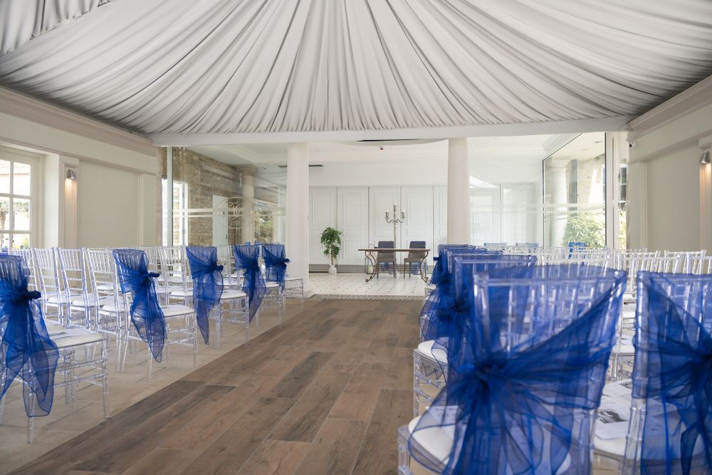 Wedding venue interior with draped fabric ceiling and blue-accented ghost chairs arranged in rows.