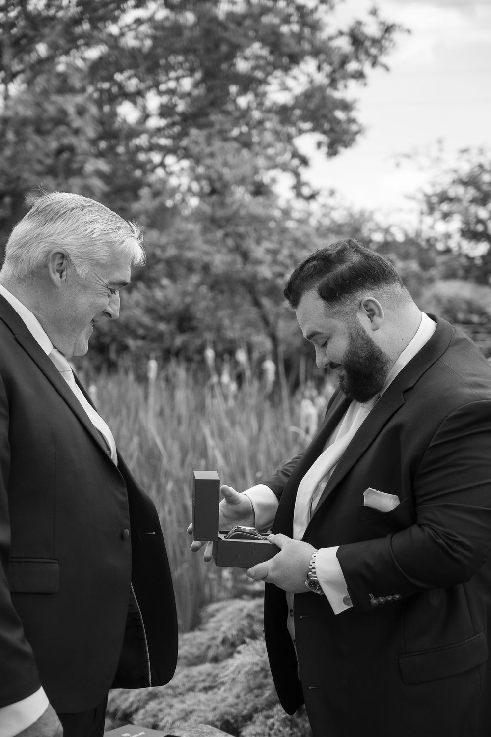 Two men in formal suits examine something together outdoors in black and white photography.