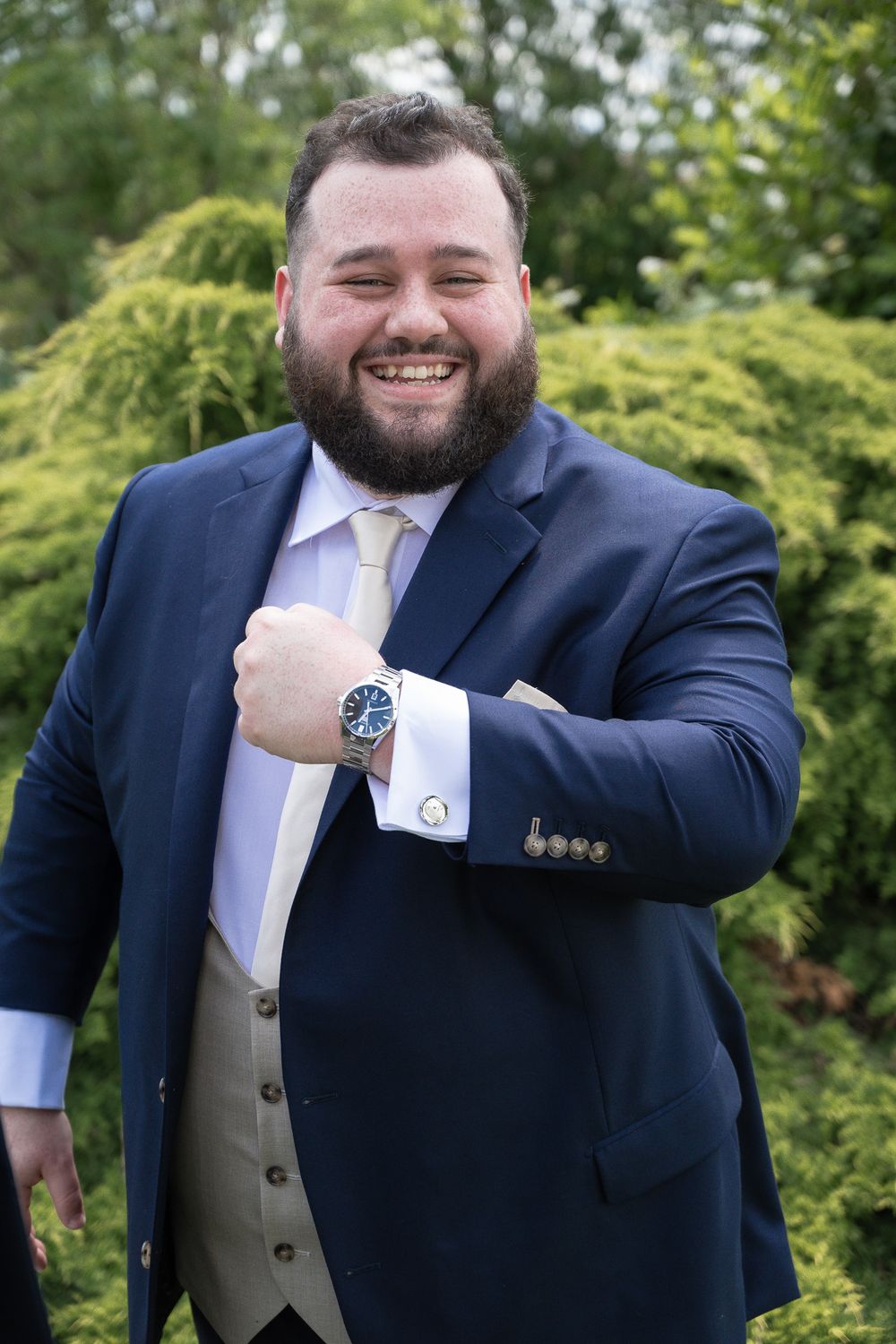 Gentleman in navy suit and tie shows off his watch while posing outdoors.