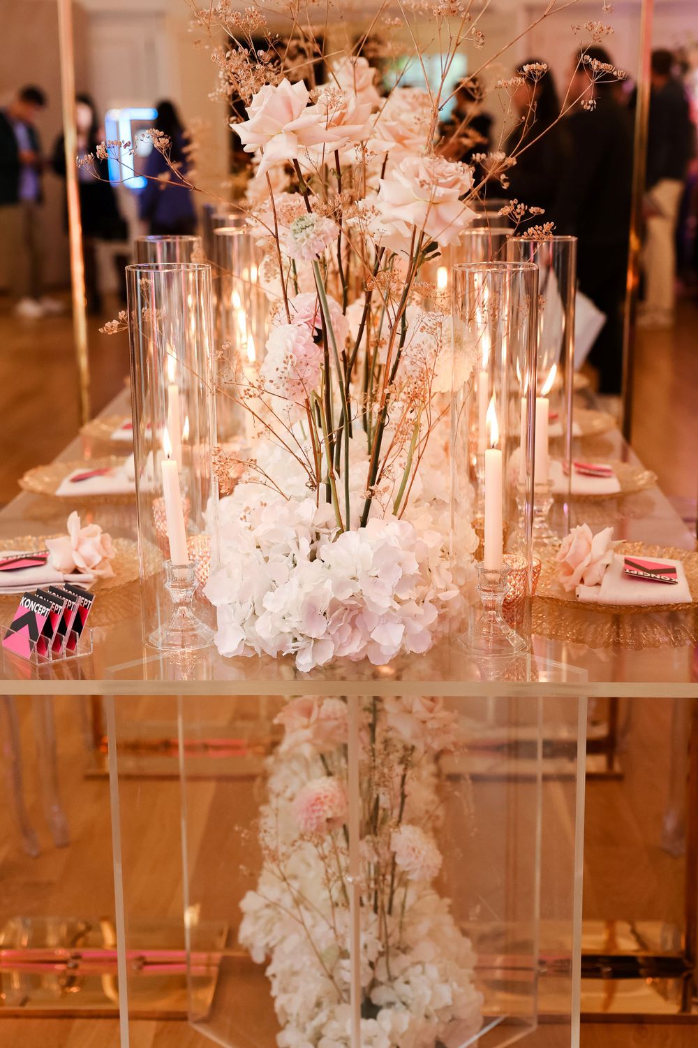 Delicate white floral centerpieces with tall glass vases on a mirrored reception table.