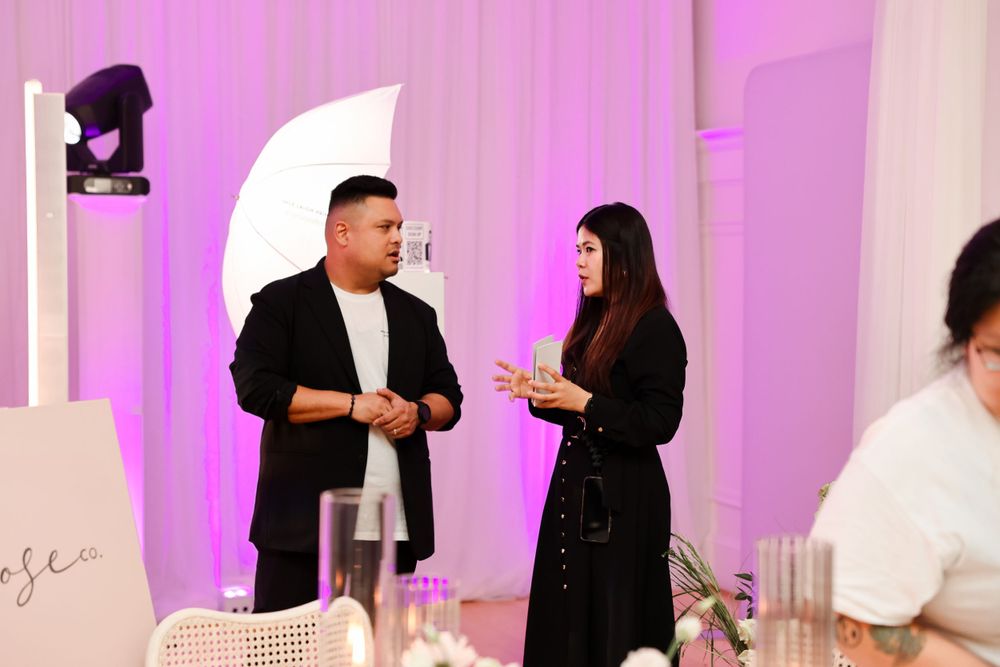 Two people in black attire having an animated conversation at an event with pink lighting and studio equipment.