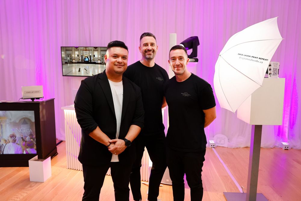 Three people in black outfits posing together in a photo studio with lighting equipment and pink accents.