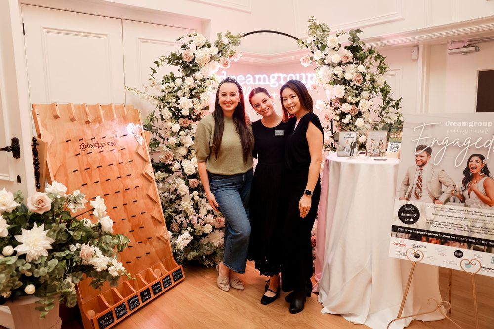 Three friends pose in front of a floral arch display at an event with decorative signage.