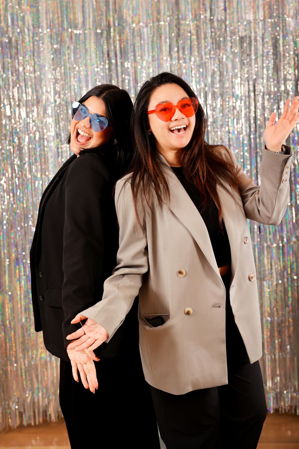 Two friends wearing colorful sunglasses pose playfully against a sparkly silver backdrop.