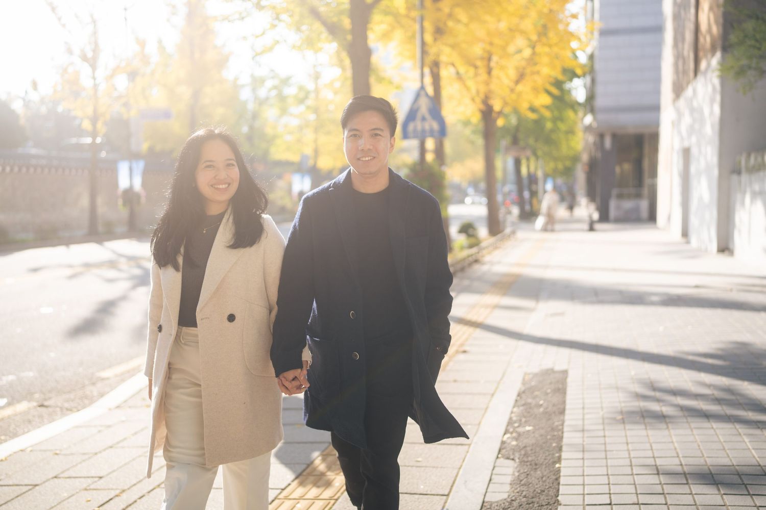 Two people walk together on an autumn sidewalk with golden yellow trees overhead and sunlight filtering through.