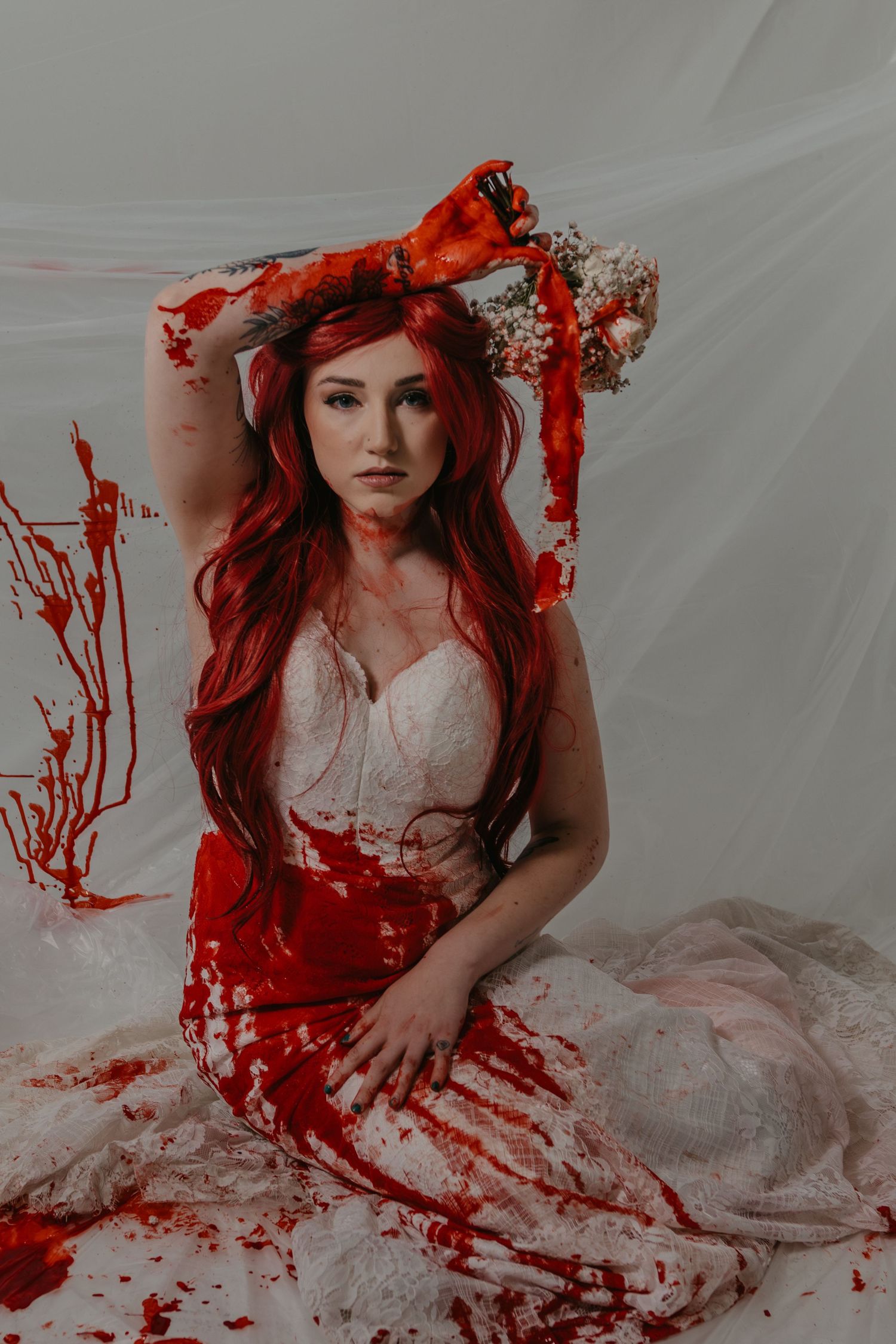 Woman in blood-splattered white wedding dress with red hair on white backdrop.