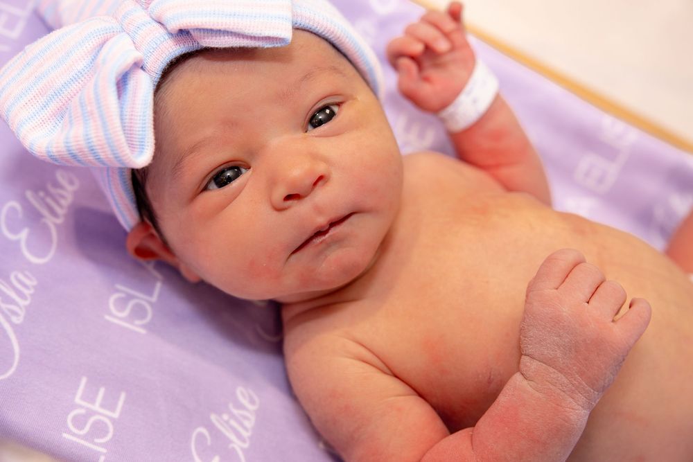 A newborn baby wearing a white bow headband lies on a purple blanket.