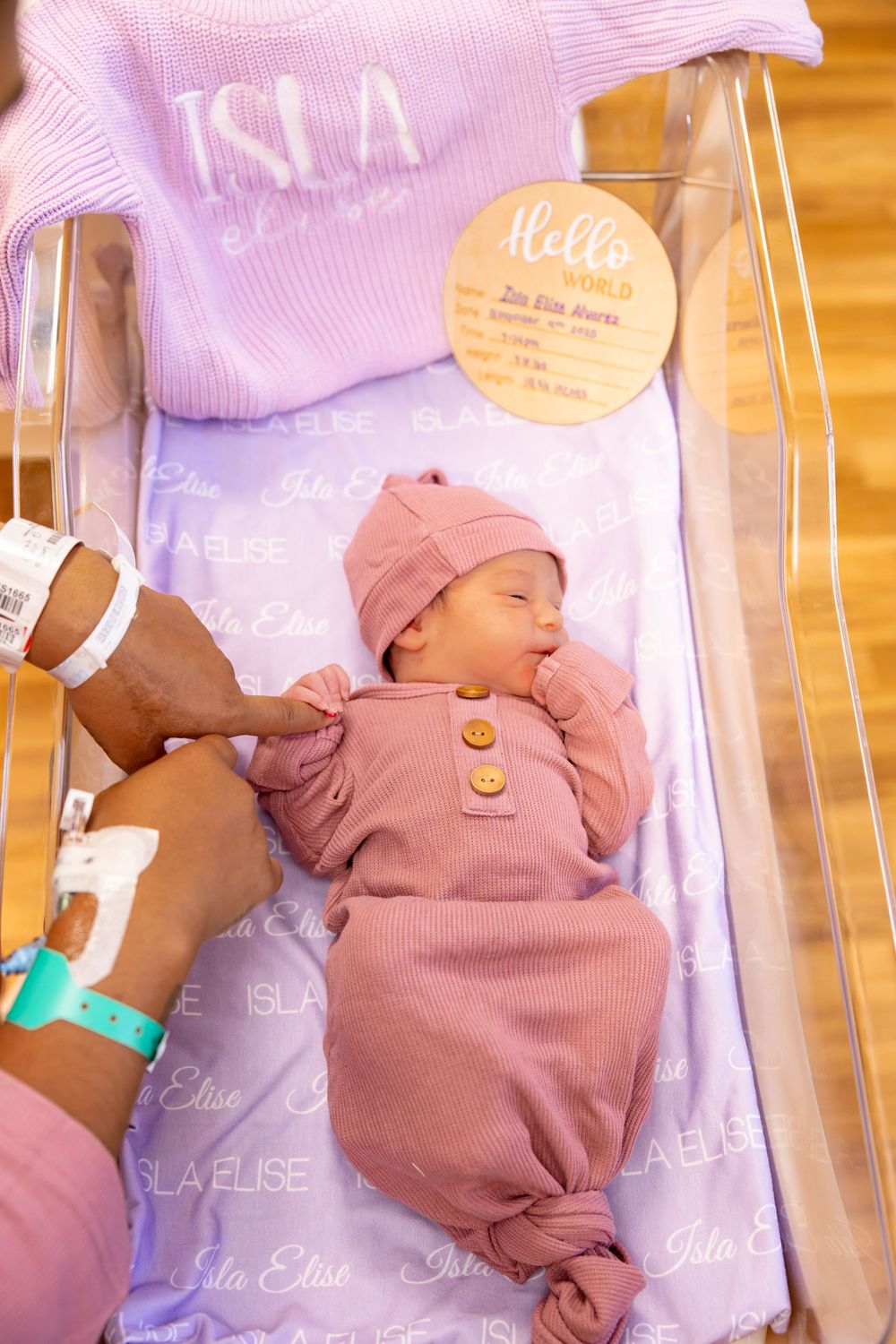 A baby wearing a pink knit hat and mauve overalls lies on a wooden surface with text overlays in soft purple tones.
