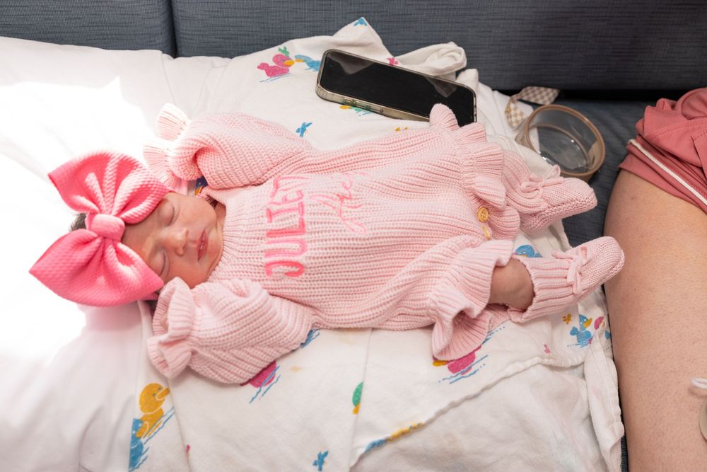 Baby wearing pink striped outfit with bow lying on white blanket with toys.