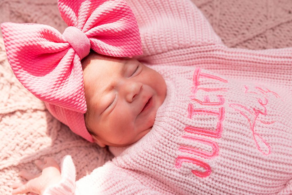 Newborn wearing a pink knit sweater and large pink bow headband sleeps peacefully on pink blanket.