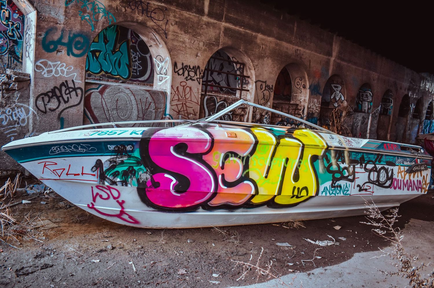 Colorful graffiti-covered speedboat abandoned under a bridge in urban setting.