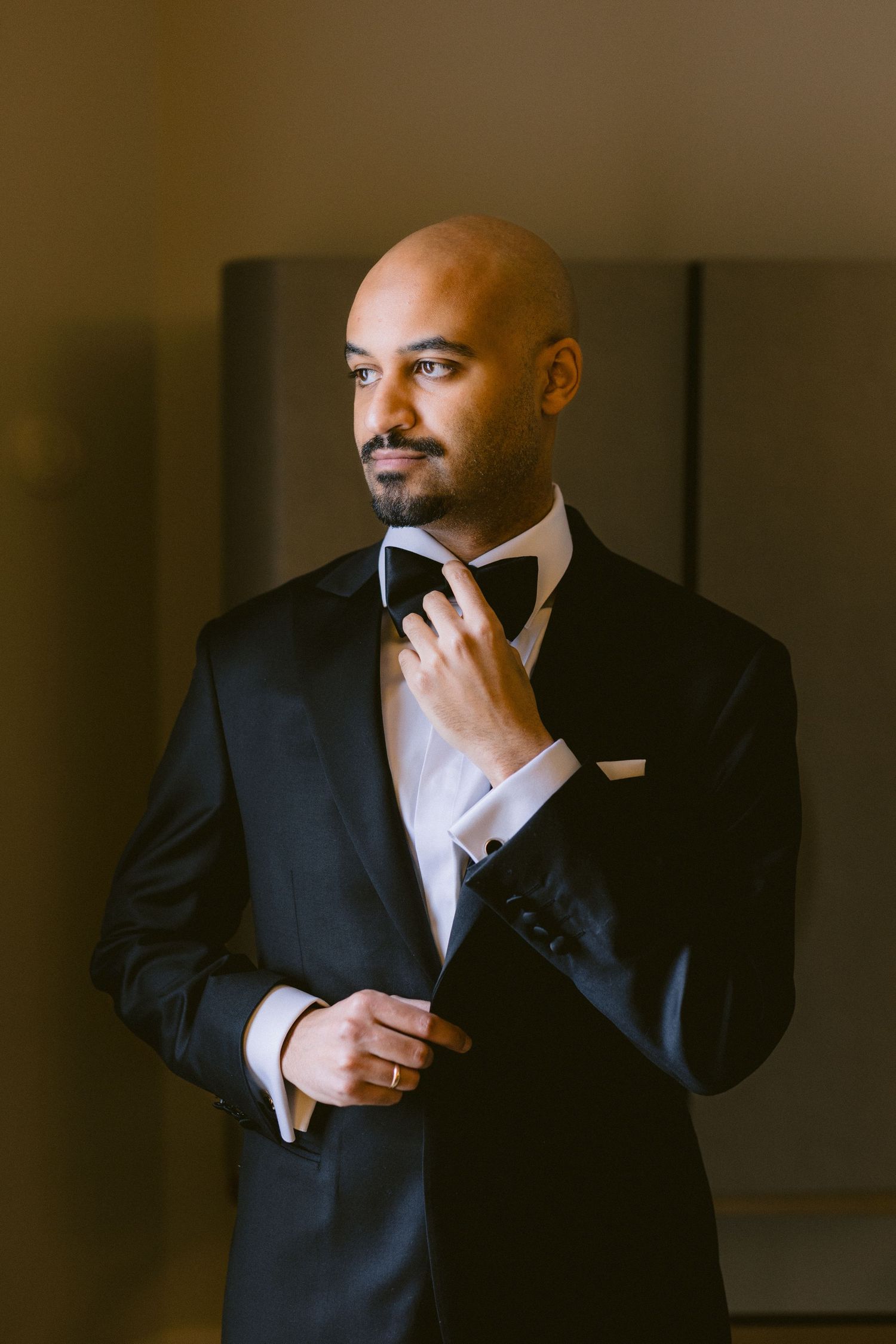 A man in a dark suit adjusts his tie while standing in a dimly lit room.