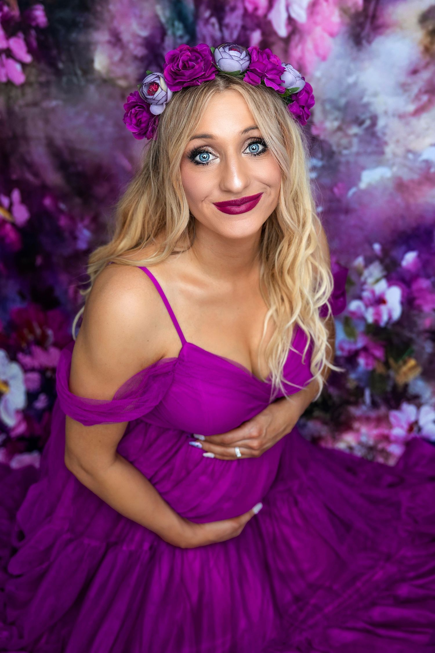 A woman in a vibrant purple gown and floral crown poses against a dreamy purple floral backdrop for a photoshoot.