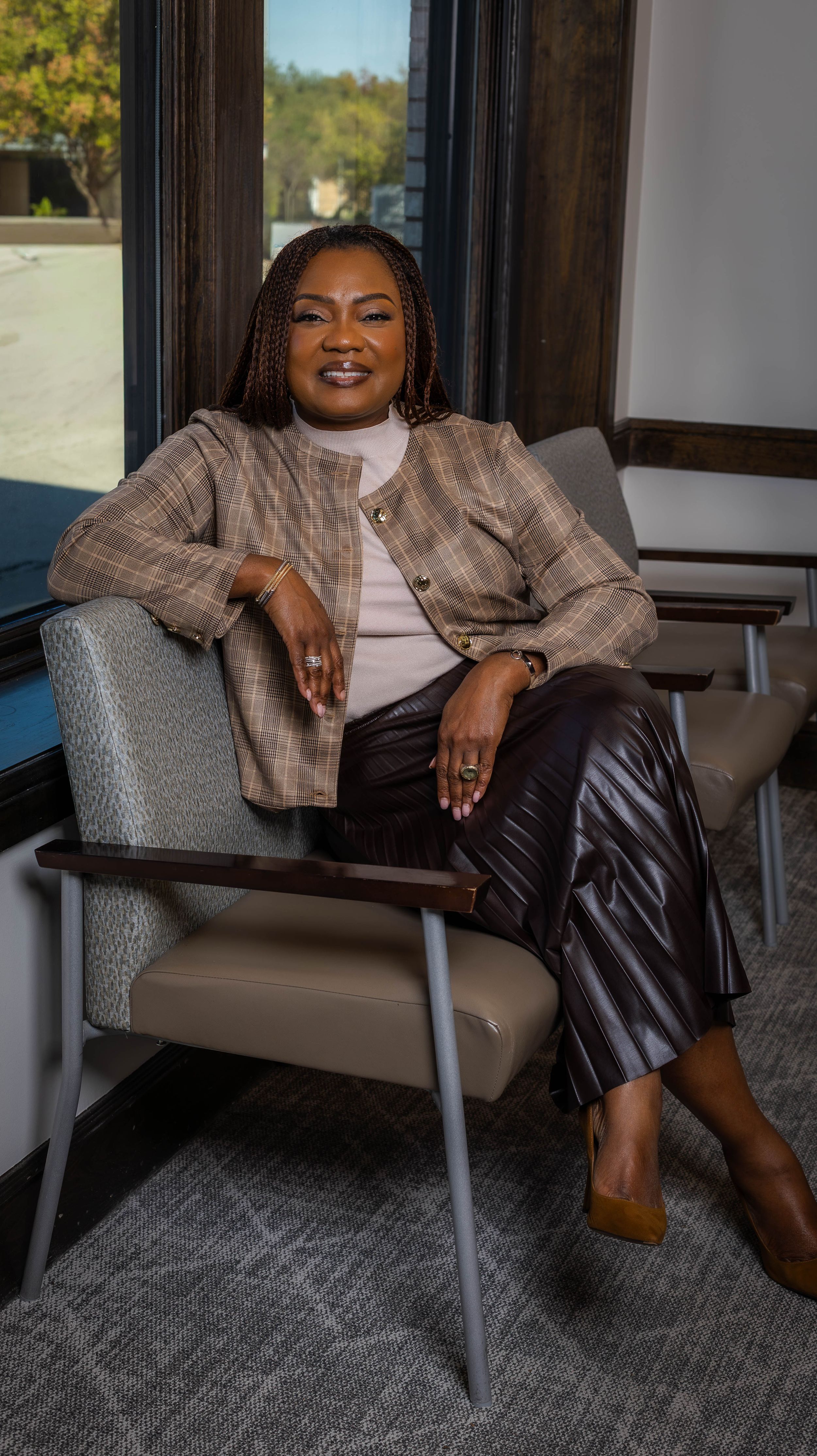 Professional seated portrait in modern office wearing plaid blazer and leather pants.