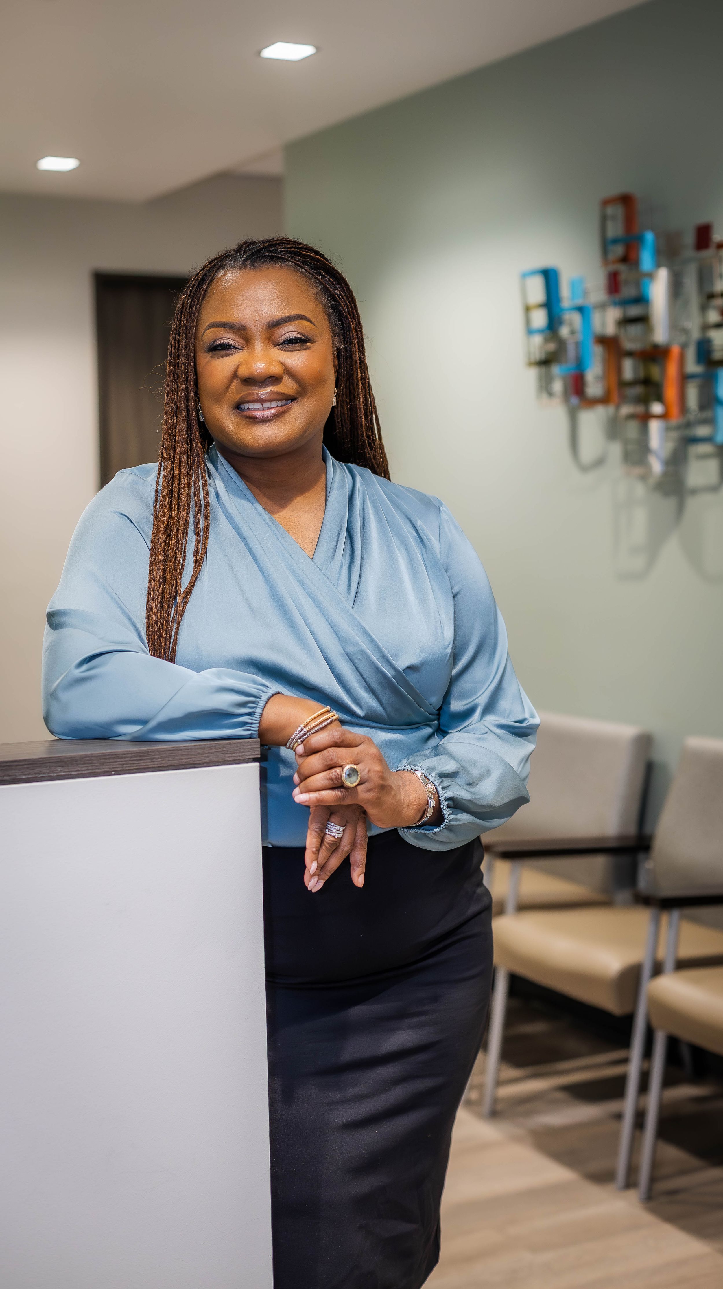 Healthcare worker in blue wrap top and black skirt stands in medical office setting.