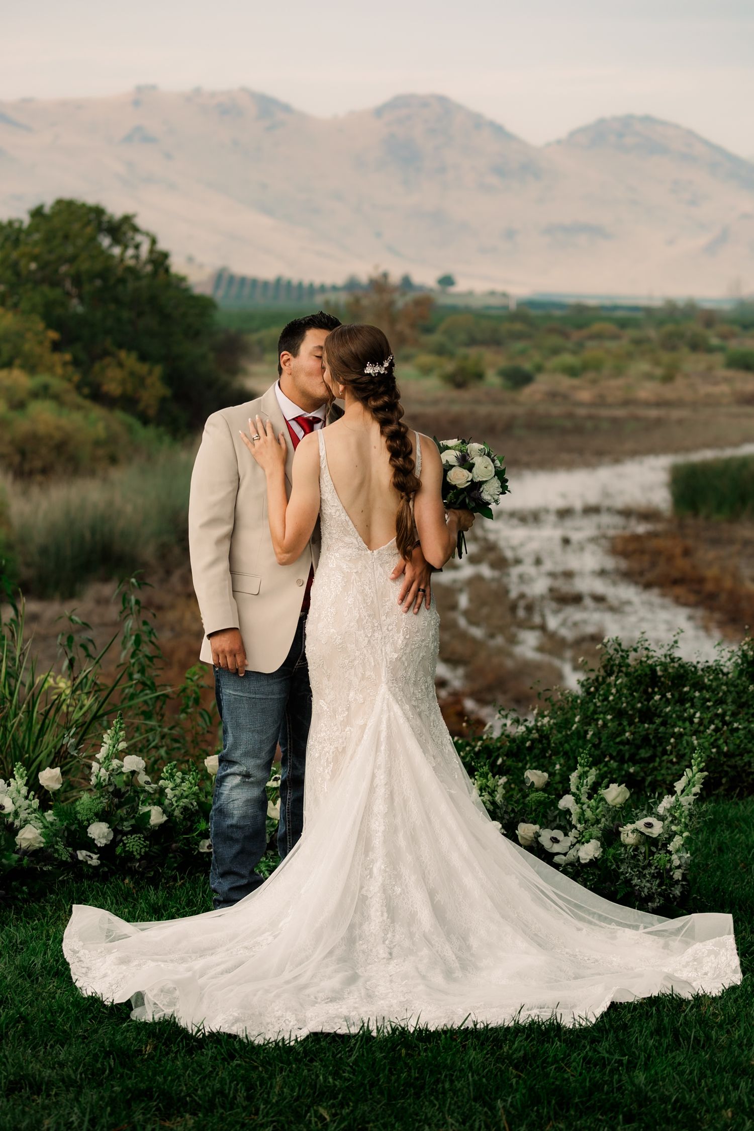 Elegant lace wedding dress with long train photographed against mountain landscape at sunset.
