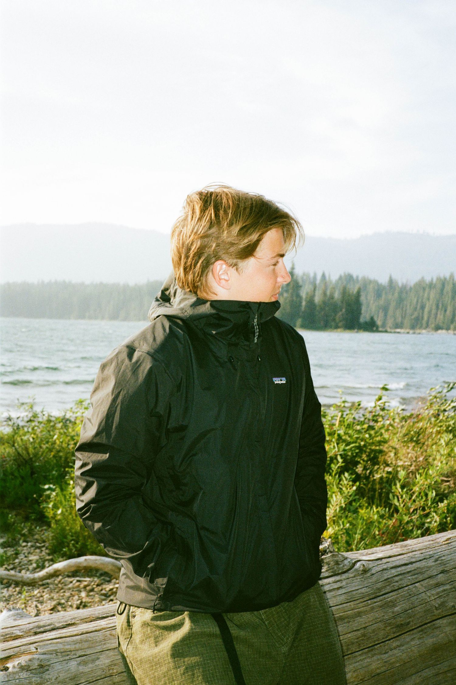Person in black jacket sitting on driftwood overlooking coastal waters.