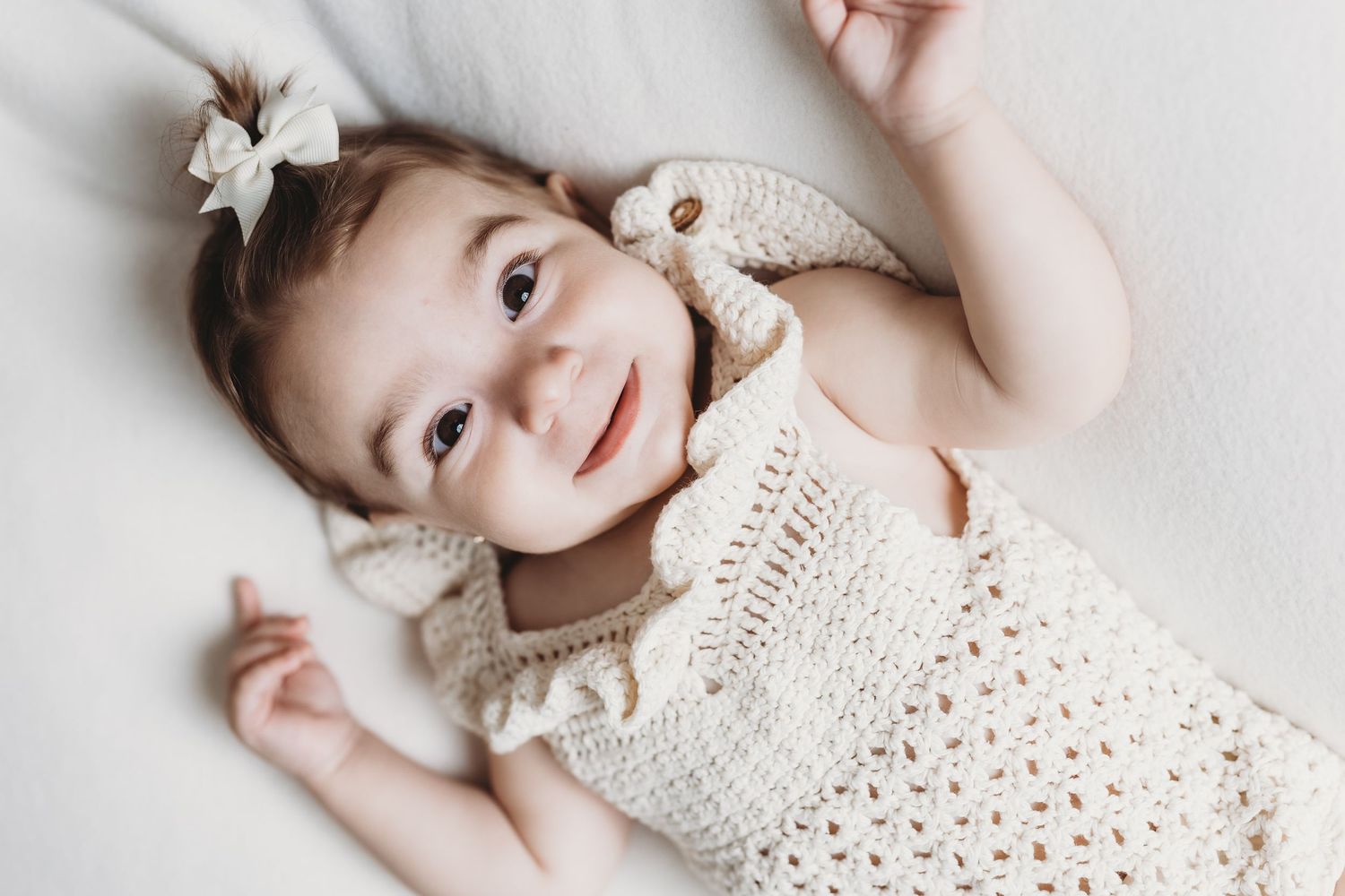 Baby wearing white crochet dress lying on white background.