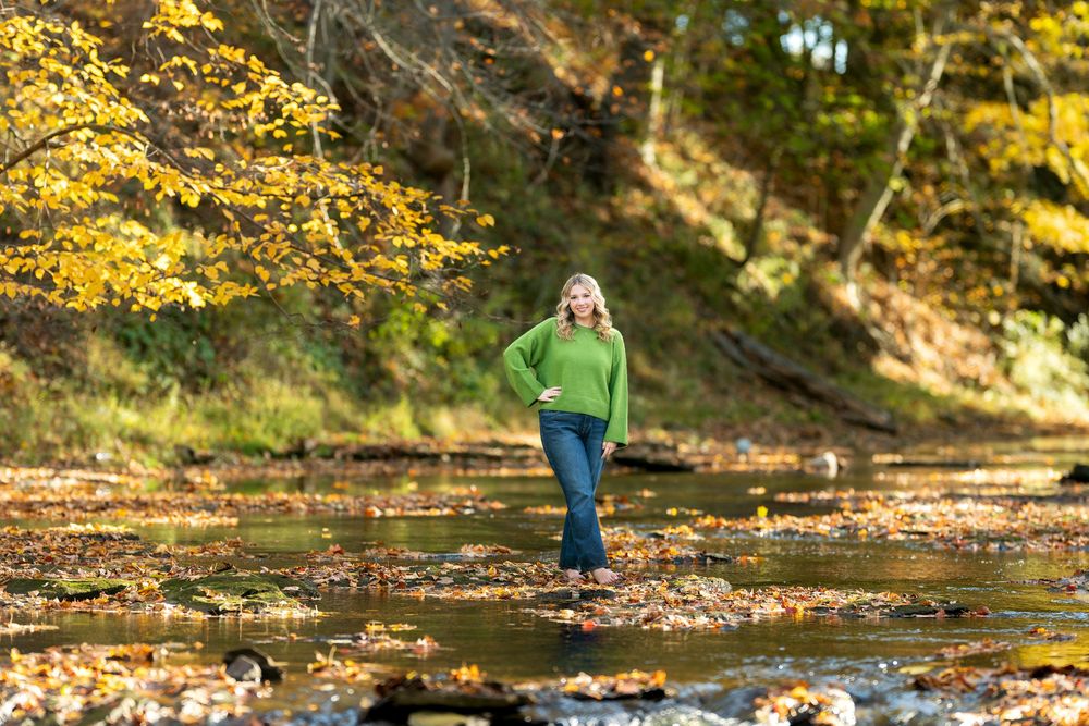 Young woman in green sweater and jeans walking through a scenic creek with golden autumn leaves.