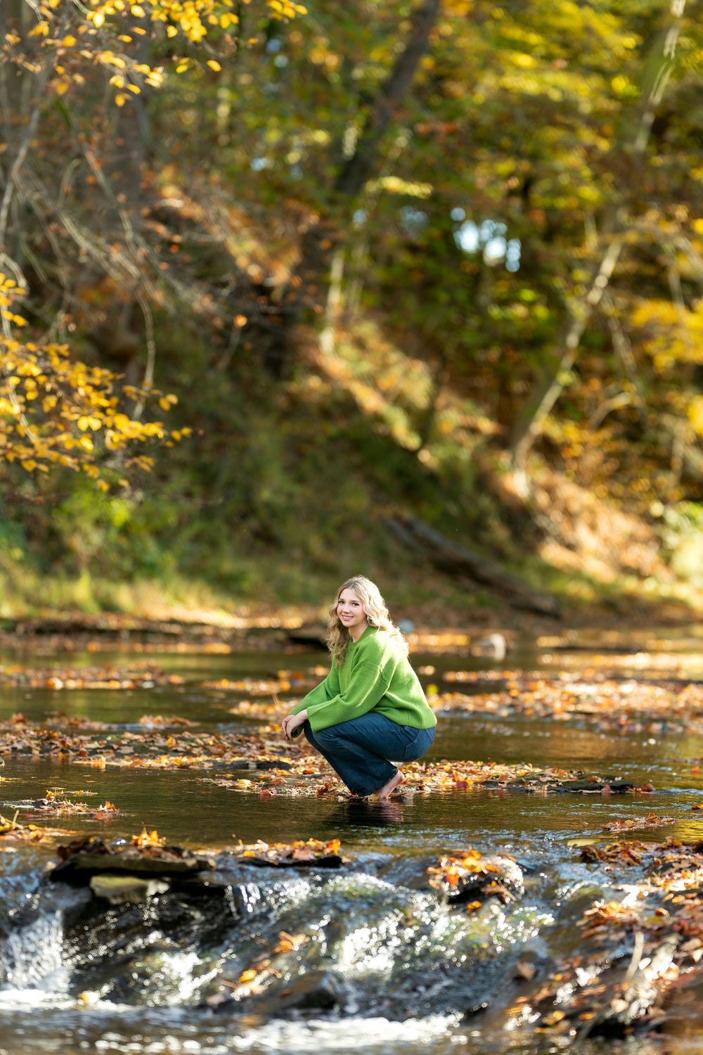 Woman in green sweater crouching in a shallow autumn creek surrounded by colorful fall foliage.