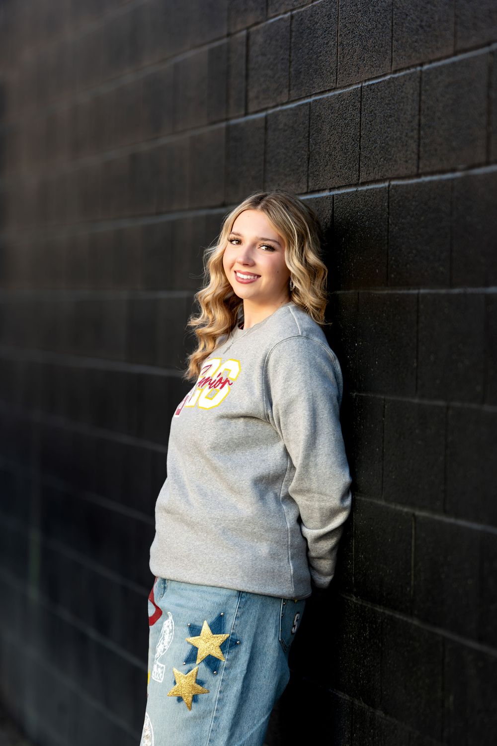 Smiling teenage girl in gray senior sweatshirt and star-print jeans leaning against a dark brick wall.