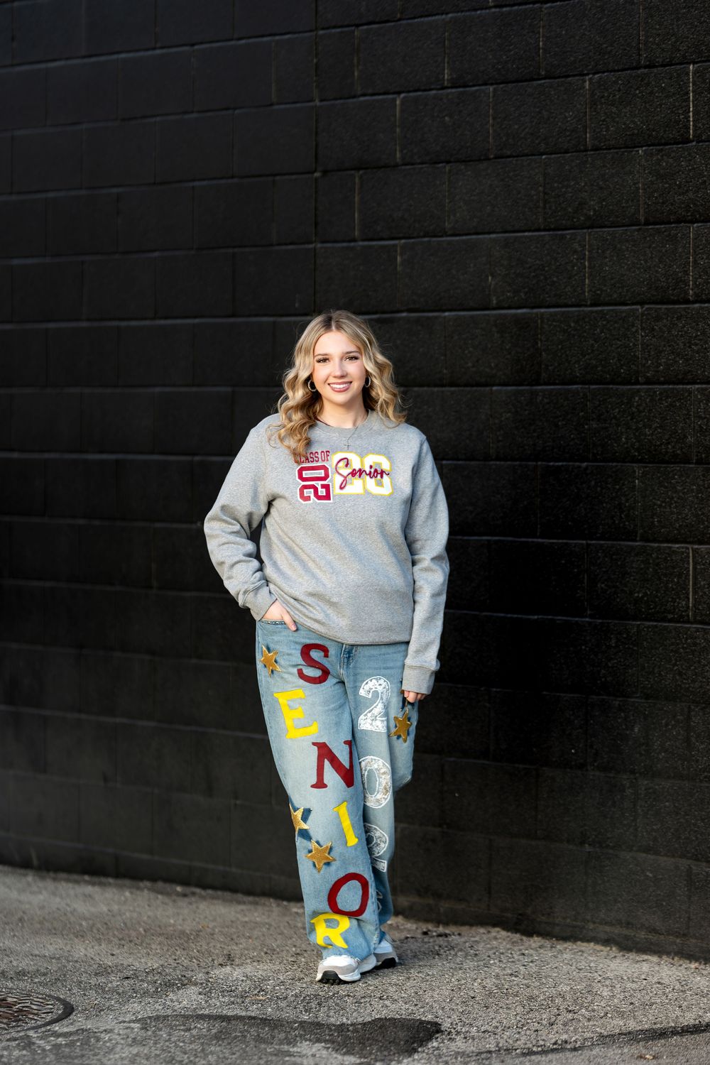 Smiling high school senior in gray sweatshirt and custom senior jeans posing against a dark brick wall.