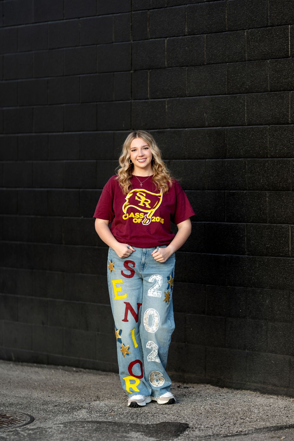 High school senior girl in SR Class of 2026 maroon tee and custom letter jeans standing on pavement.