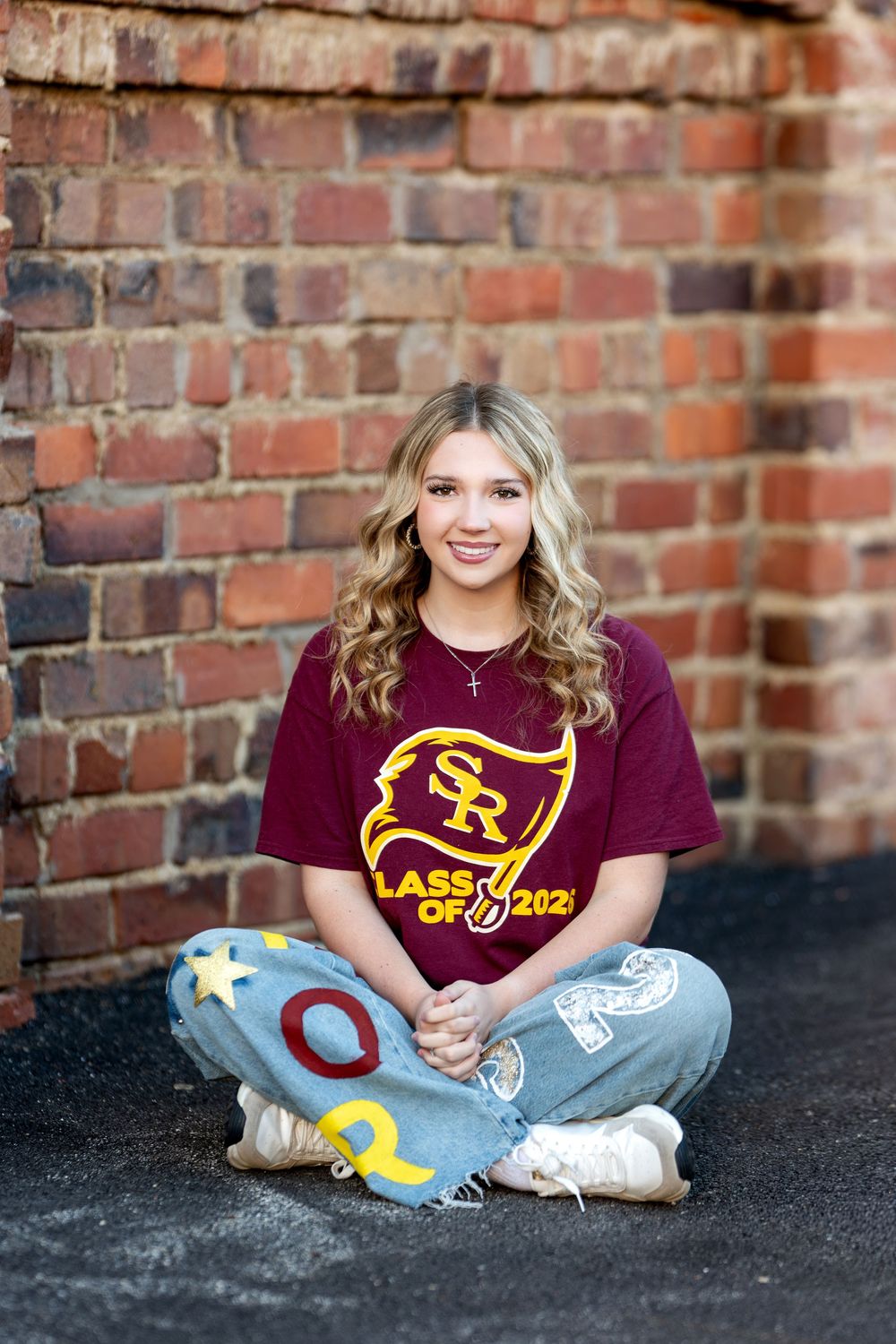 High school senior girl sitting cross-legged in SR Class of 2026 shirt and custom jeans by brick wall.