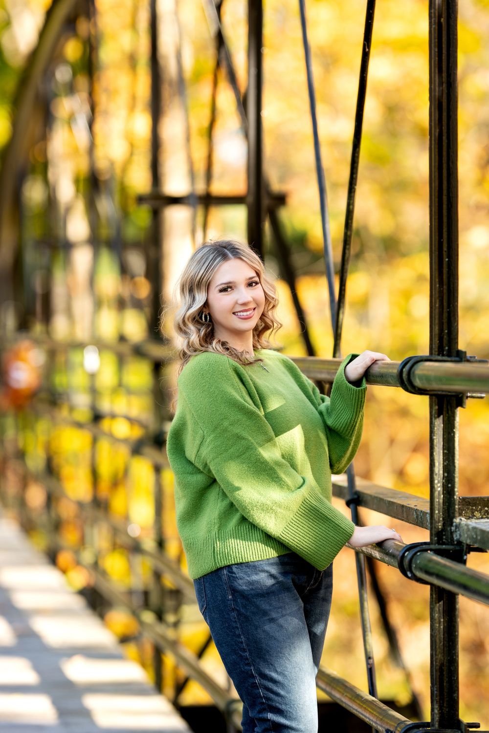 Blonde woman in green sweater smiling on iron bridge surrounded by autumn trees.