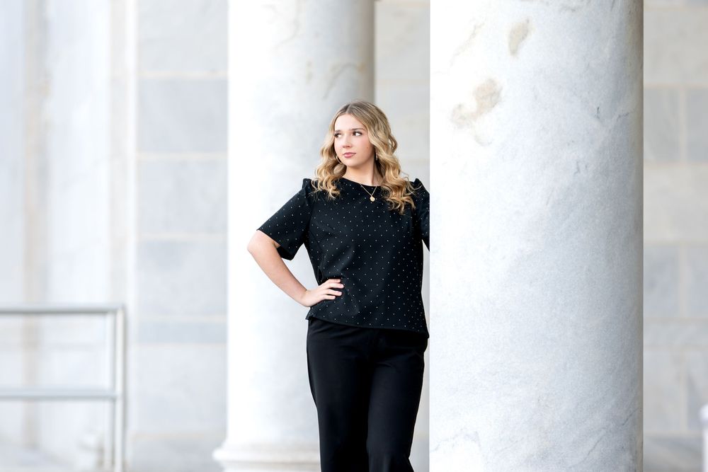 Young blonde woman in black polka dot top and wide-leg pants posing beside white marble column.