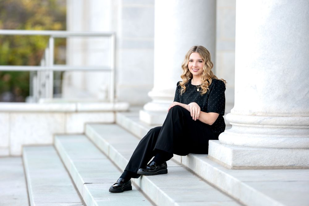 Smiling senior girl in black outfit sitting casually on marble steps beside tall white columns.