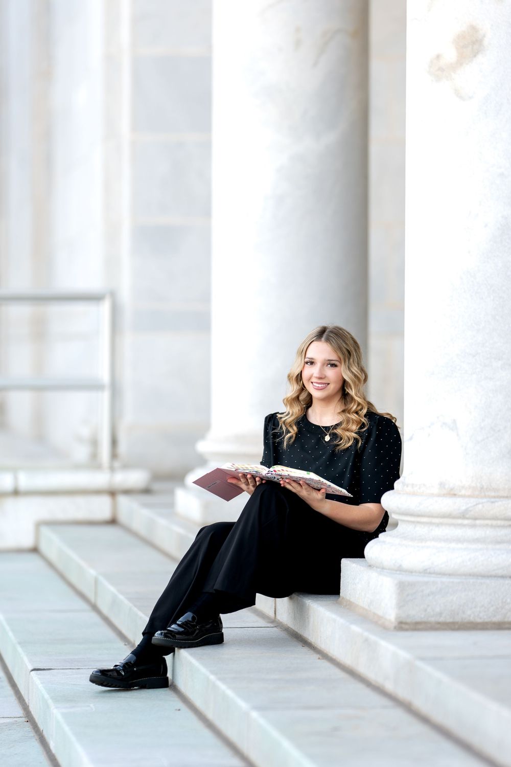 Smiling blonde senior girl reading a book on marble steps surrounded by tall white columns.