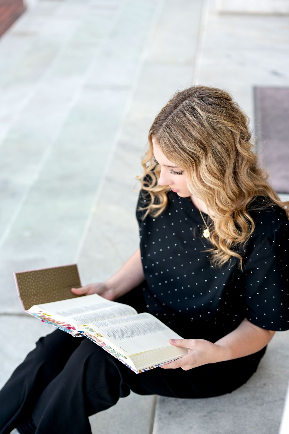 Overhead view of blonde teen girl in black polka dot dress reading an open book on stone steps.