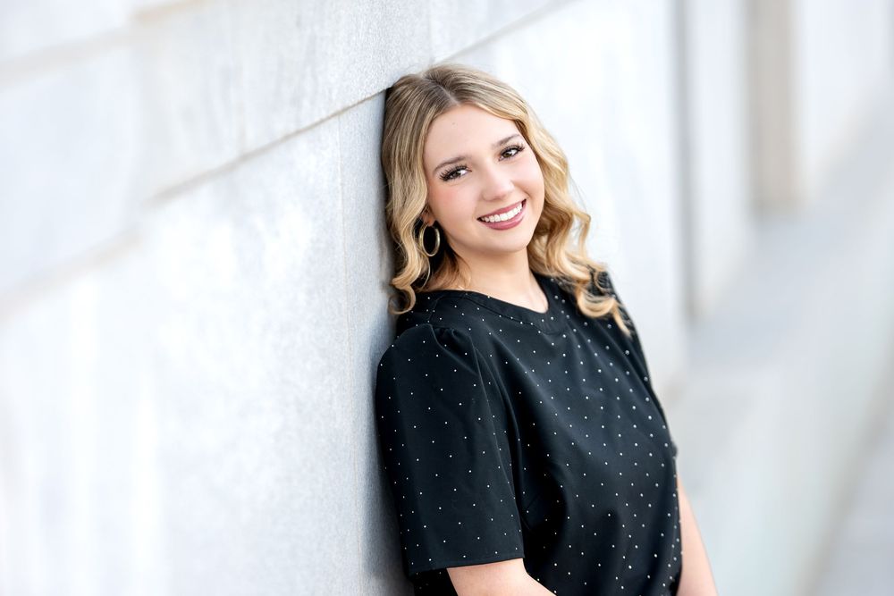Blonde woman in black polka dot top smiling over shoulder against marble wall.