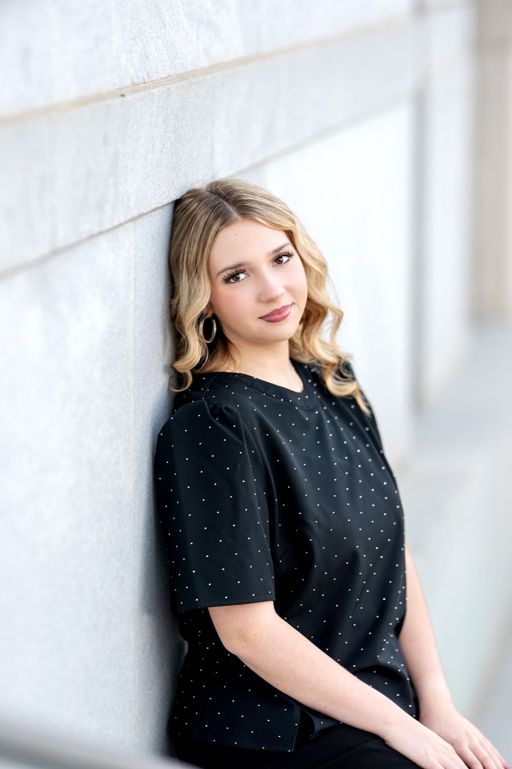 Young blonde woman in black dotted top leaning against marble wall with subtle smile.