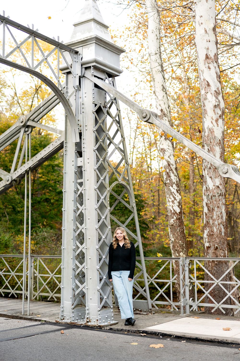 Blonde teen girl in black sweater and light jeans posing by historic iron bridge tower amid fall foliage.