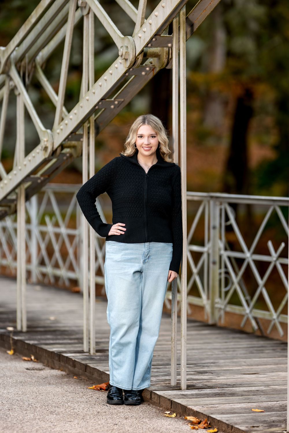 Young woman in black zip-up sweater and light jeans posing on vintage iron bridge for senior photos.