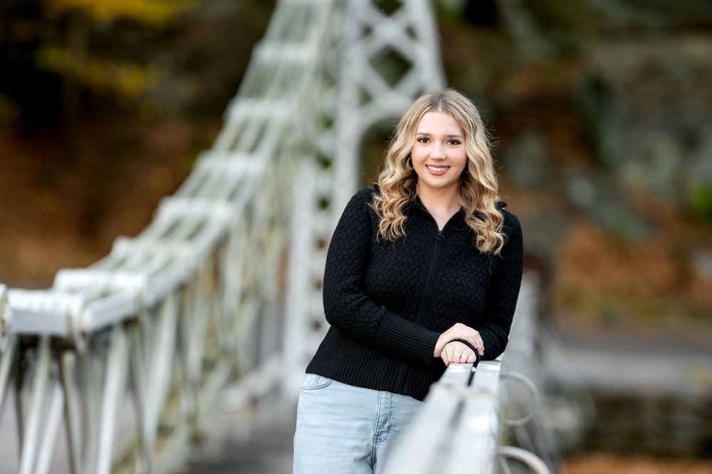 Young blonde woman in black sweater leaning on iron bridge railing surrounded by autumn foliage.