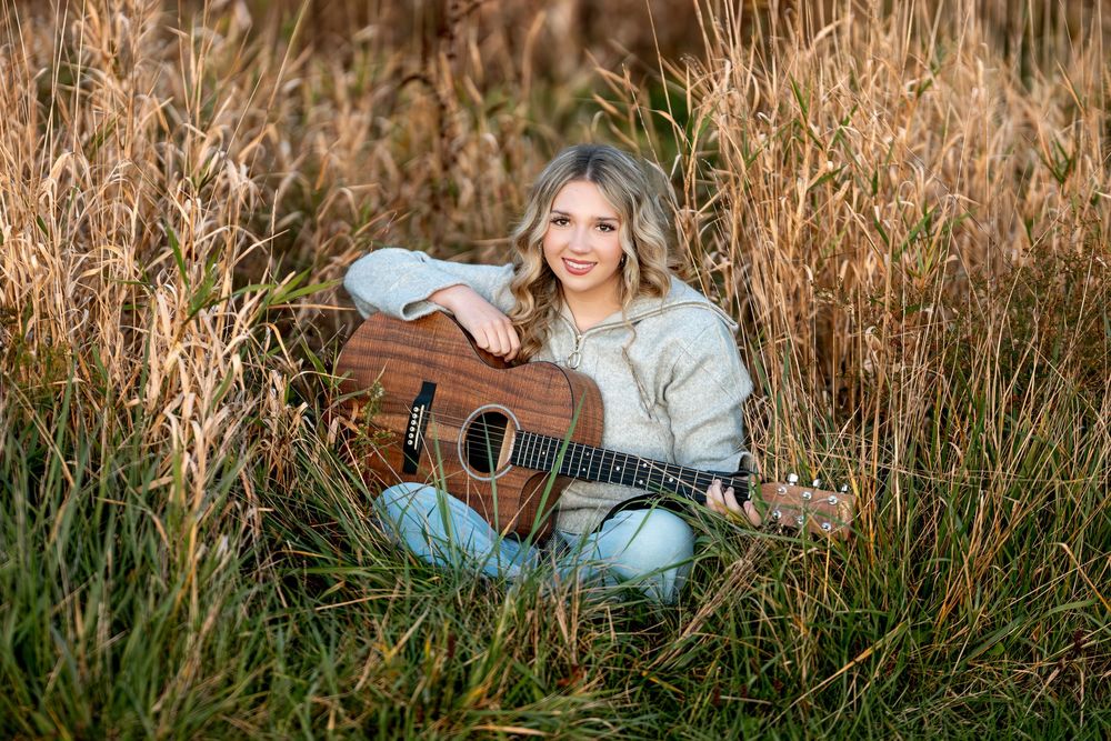Smiling blonde teen sitting in tall golden grass field hugging acoustic guitar in warm fall light.