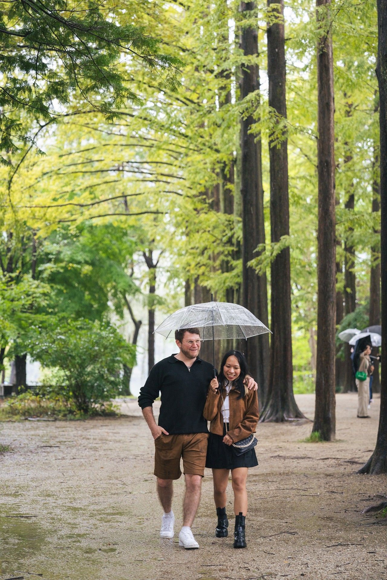 A couple walks through a scenic tree-lined path holding umbrellas on an autumn day.