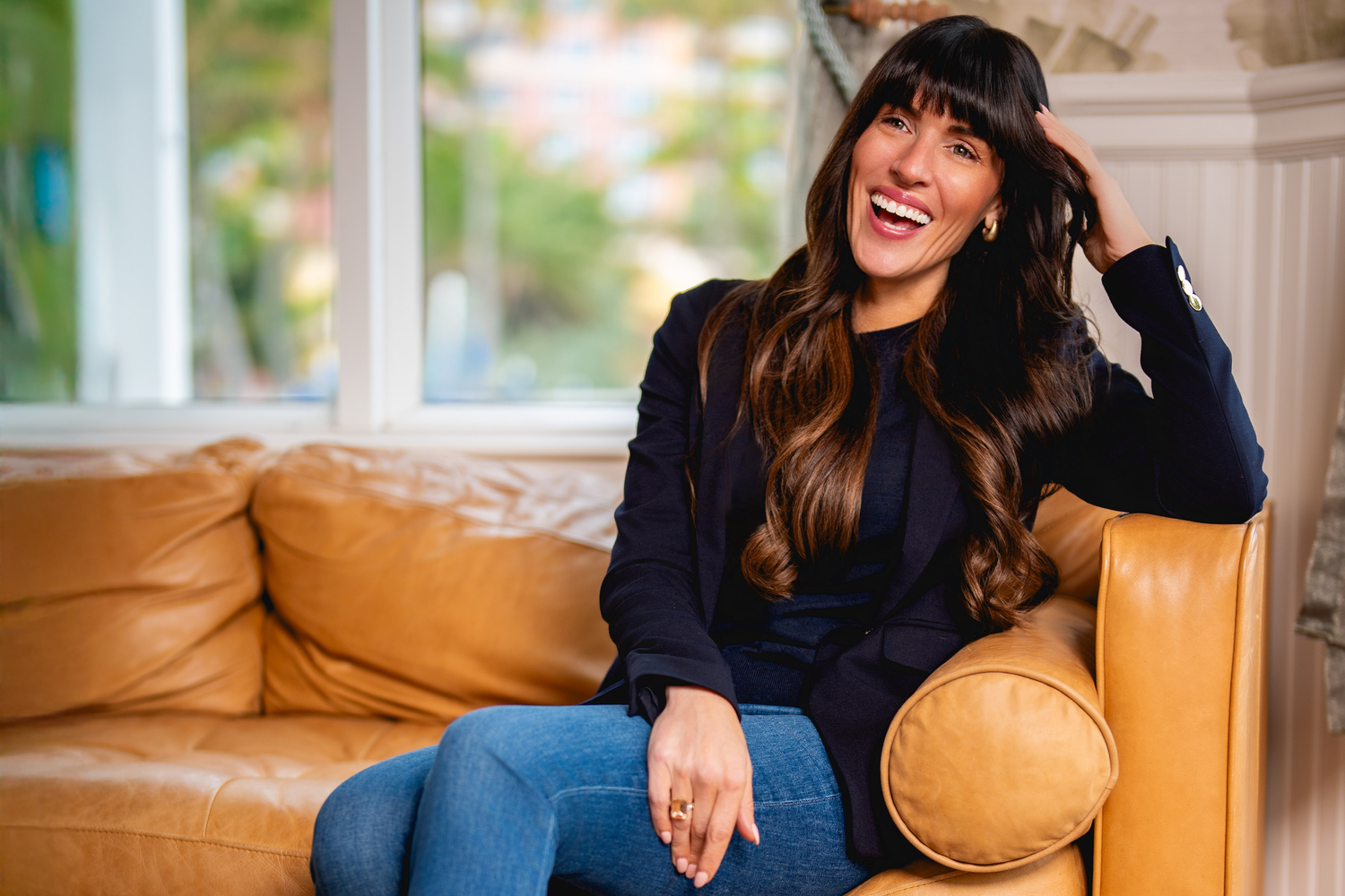 A person laughs joyfully while sitting on a tan leather couch in a bright interior setting.