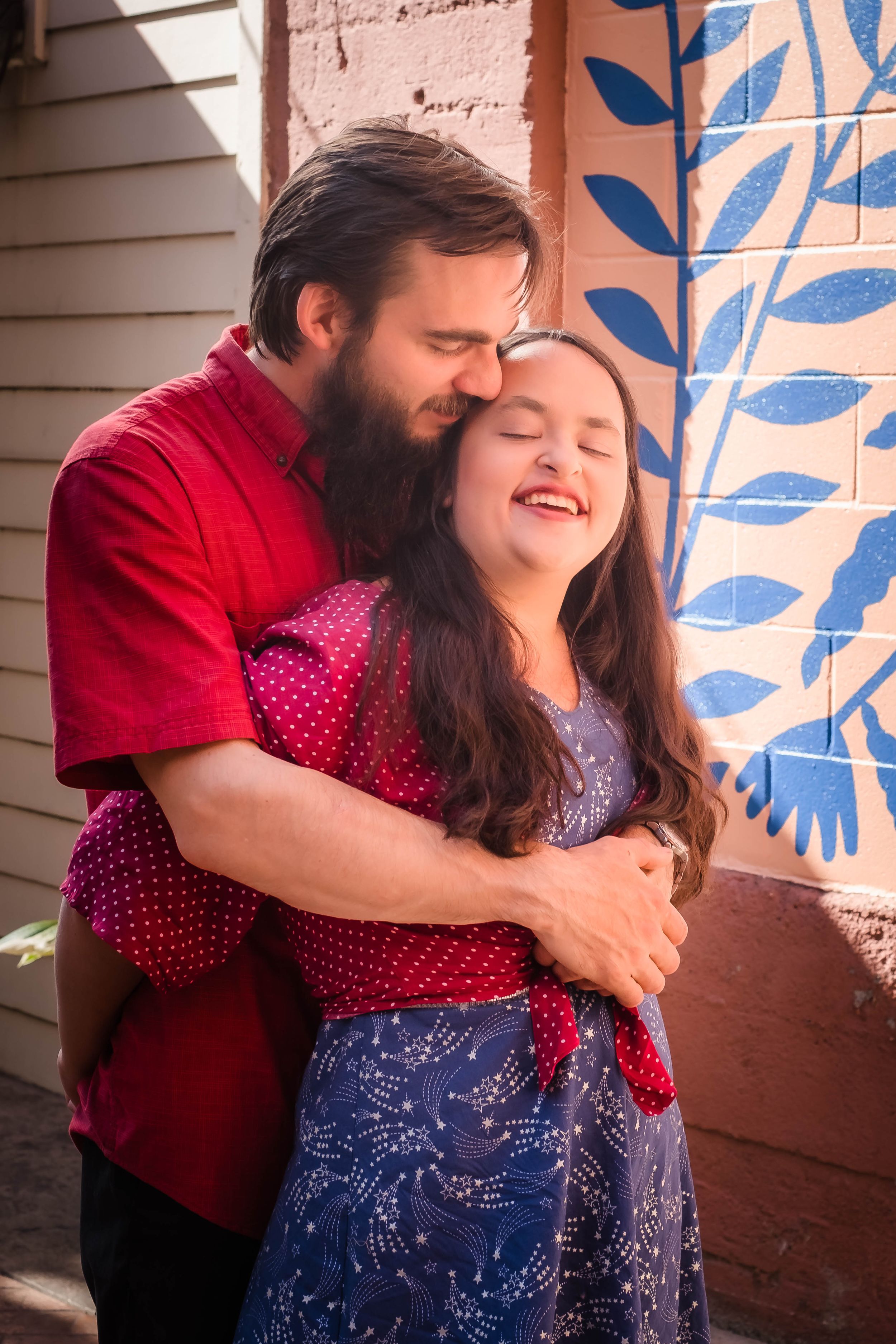 A couple in red and blue outfits share a tender embrace while standing on a porch with decorative blue leaf patterns.