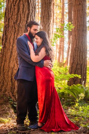 A couple embraces romantically in a sunlit forest setting with the woman wearing a flowing red velvet dress.