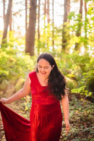 A person in a flowing red velvet dress twirls joyfully in a sunlit forest setting during golden hour.