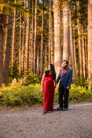 A couple in a red dress and dark suit share intimate moments among tall pine trees in a forest setting at sunset.
