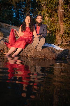 Couple in red dress sitting on rocky riverbank with water reflections at sunset.