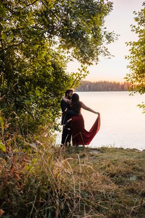 A romantic couple sits together on a grassy cliff overlooking a serene lake at golden hour.