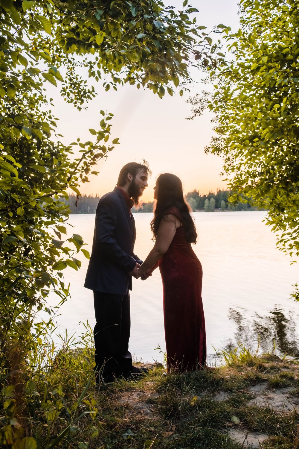 A silhouetted couple shares a tender moment by a serene lake at sunset framed by overhanging tree branches.
