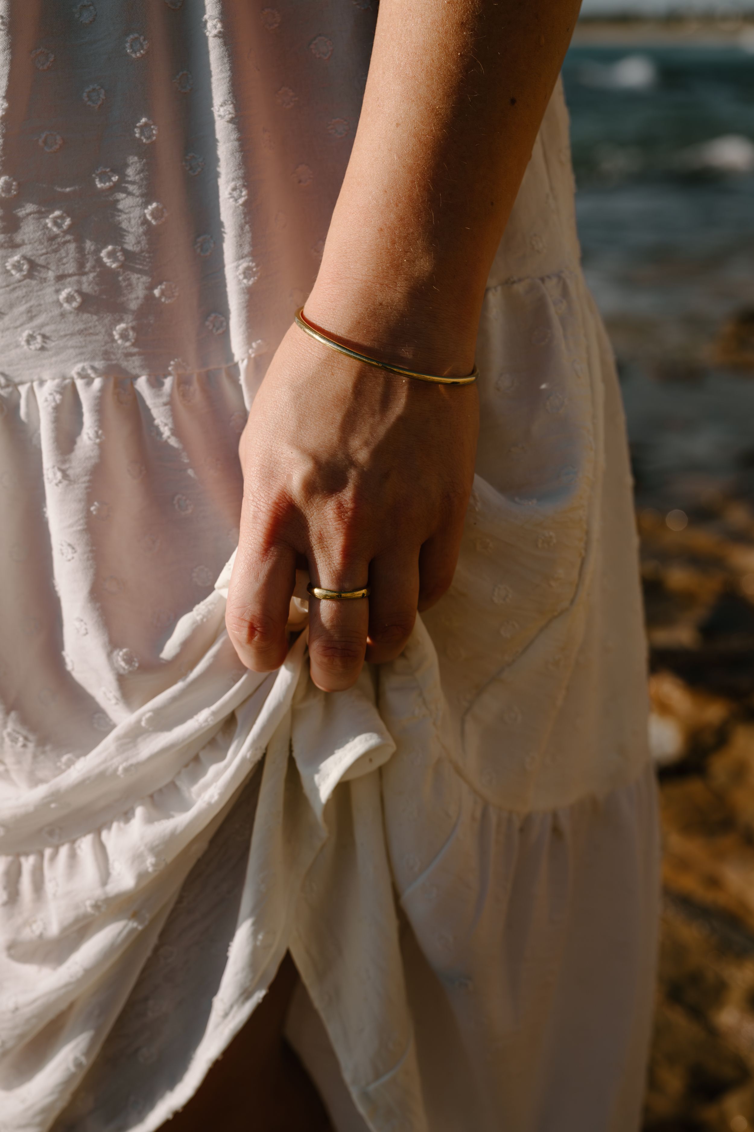 A delicate hand wearing rings and a thin bracelet holds the fabric of a flowing white dress against a natural backdrop.