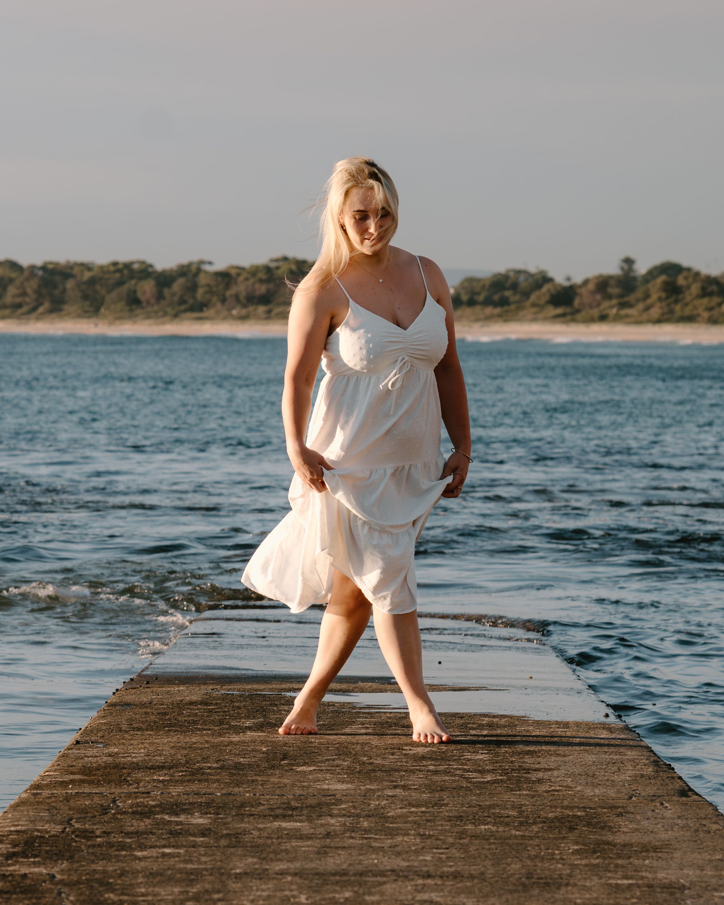 Person in a flowing white sundress walks barefoot on a wooden pier by the ocean at sunset.